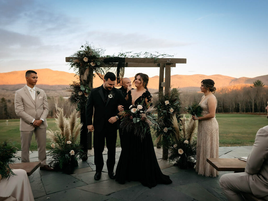couple in black formalwear in front of arbor with mountains and sky in background